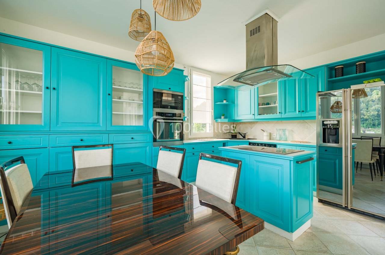 Cuisine moderne avec armoires turquoise, table en bois foncé, électroménagers intégrés et suspensions en osier.