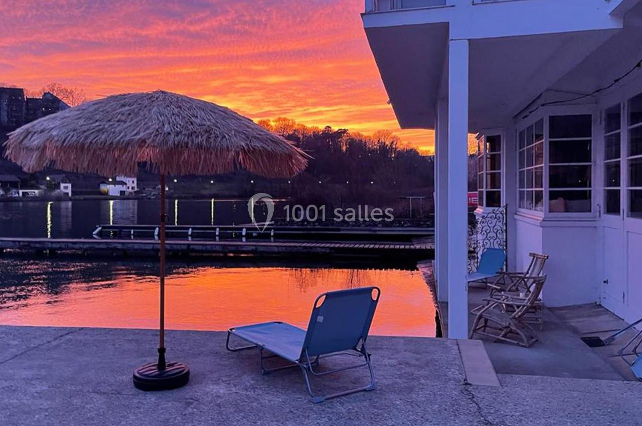 Terrasse au bord d'un lac au coucher du soleil, avec un parasol en paille, une chaise longue et des chaises en bois.