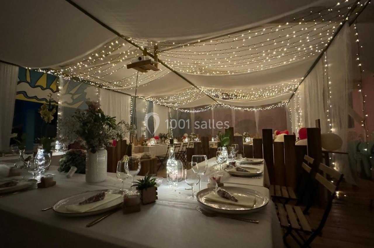 Salle de réception décorée avec des guirlandes lumineuses suspendues, tables dressées et ambiance chaleureuse.
