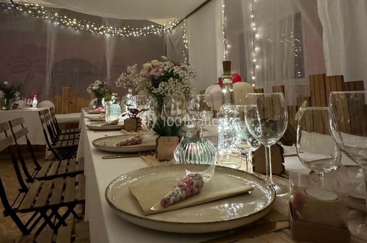 Table décorée pour un dîner festif avec vaisselle élégante, fleurs, bougies et guirlandes lumineuses en arrière-plan.