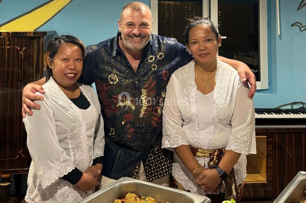 Un homme souriant pose avec deux femmes en tenue traditionnelle devant des plats de nourriture dans un intérieur coloré.