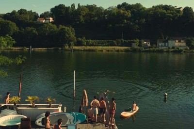 Tables et chaises en bois disposées en extérieur près d'une rivière, avec parasols et végétation en arrière-plan.