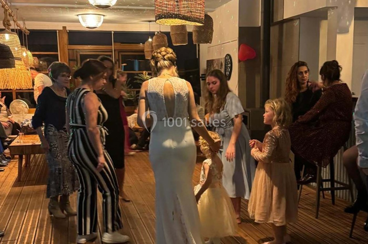 Une femme en robe blanche discute avec des enfants dans une salle éclairée, entourée de plusieurs adultes.