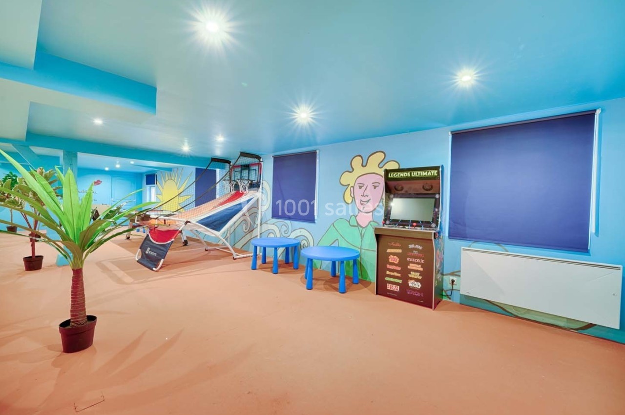 Salle colorée avec murs peints, hamac, tables bleues, borne d'arcade et plantes décoratives.