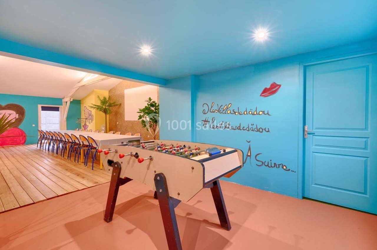 Salle colorée avec baby-foot au premier plan, grande table en bois au fond et décorations murales artistiques.