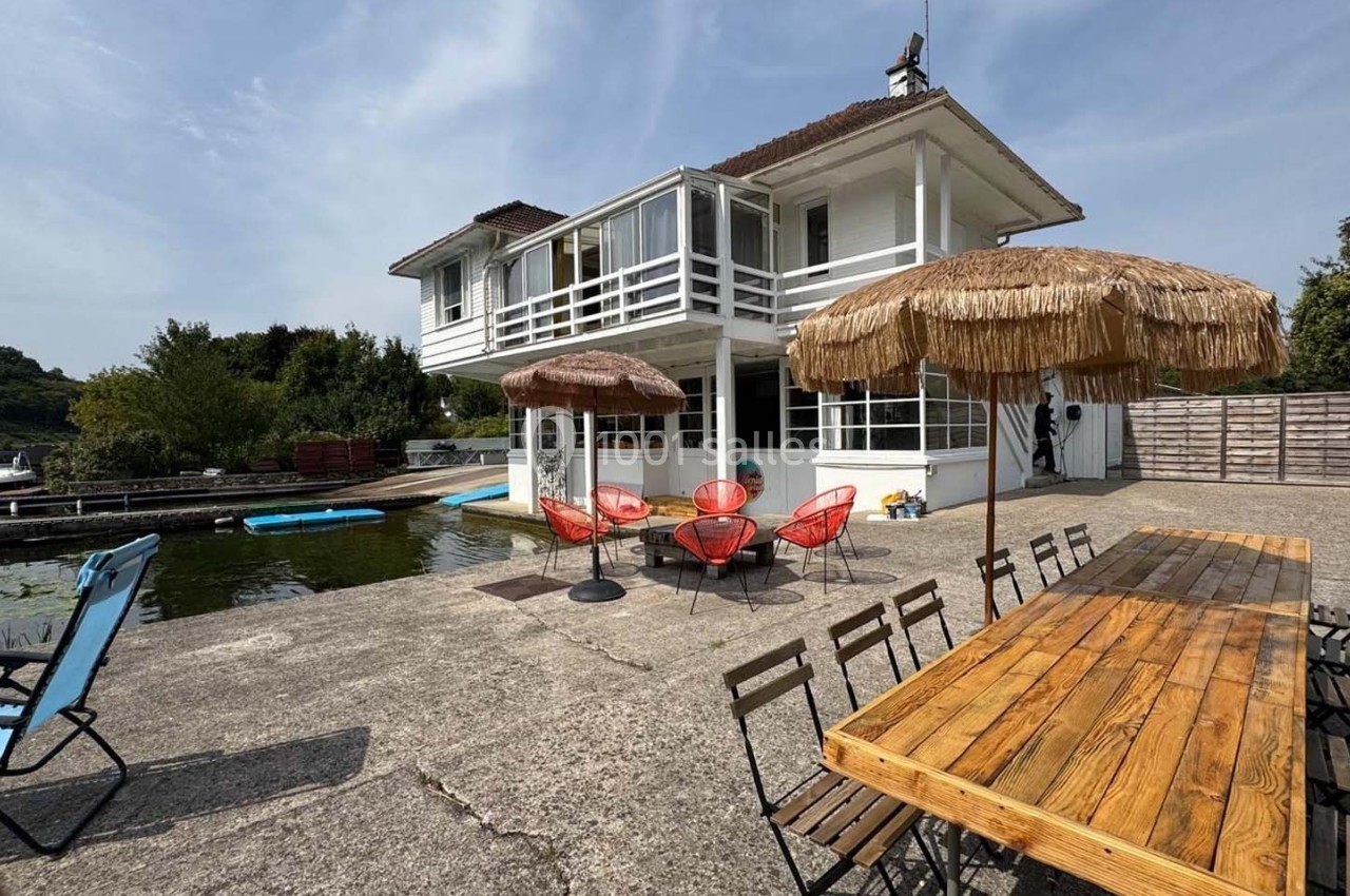 Maison au bord d’un lac avec terrasse aménagée, chaises, parasols en paille, et table en bois près d’un ponton.