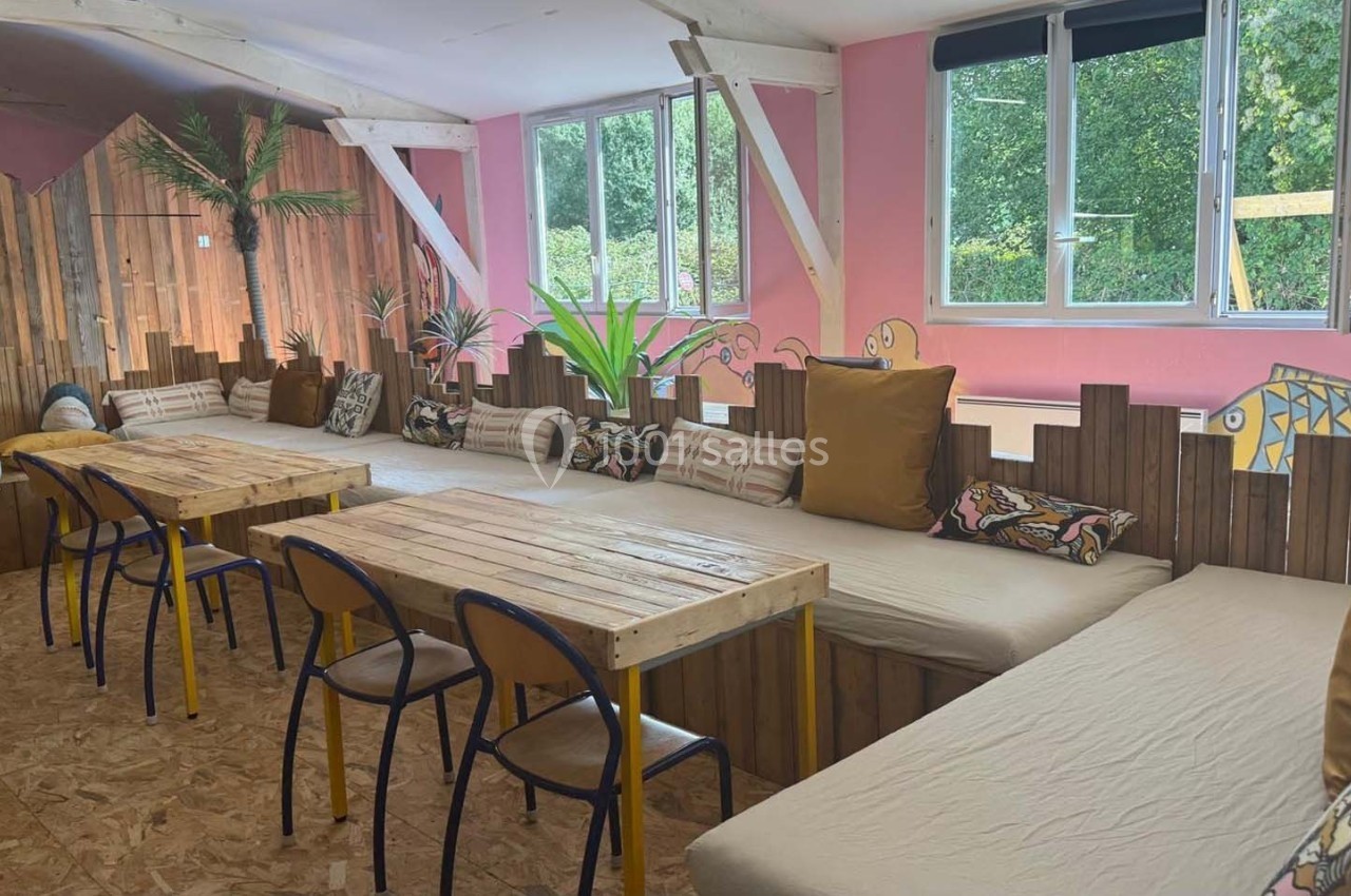 Salle lumineuse avec tables en bois, chaises colorées, banquettes garnies de coussins et décoration végétale.