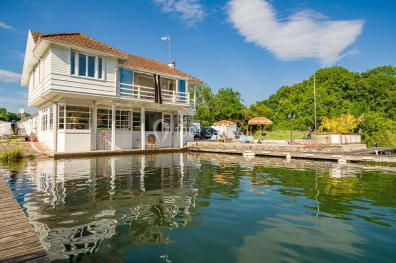 Maison blanche à deux étages au bord d'un plan d'eau, entourée de végétation sous un ciel bleu.