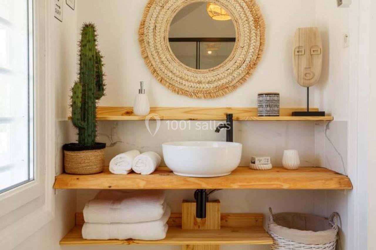 Salle de bain avec lavabo blanc, miroir rond en rotin, cactus, objets décoratifs et étagères en bois clair.