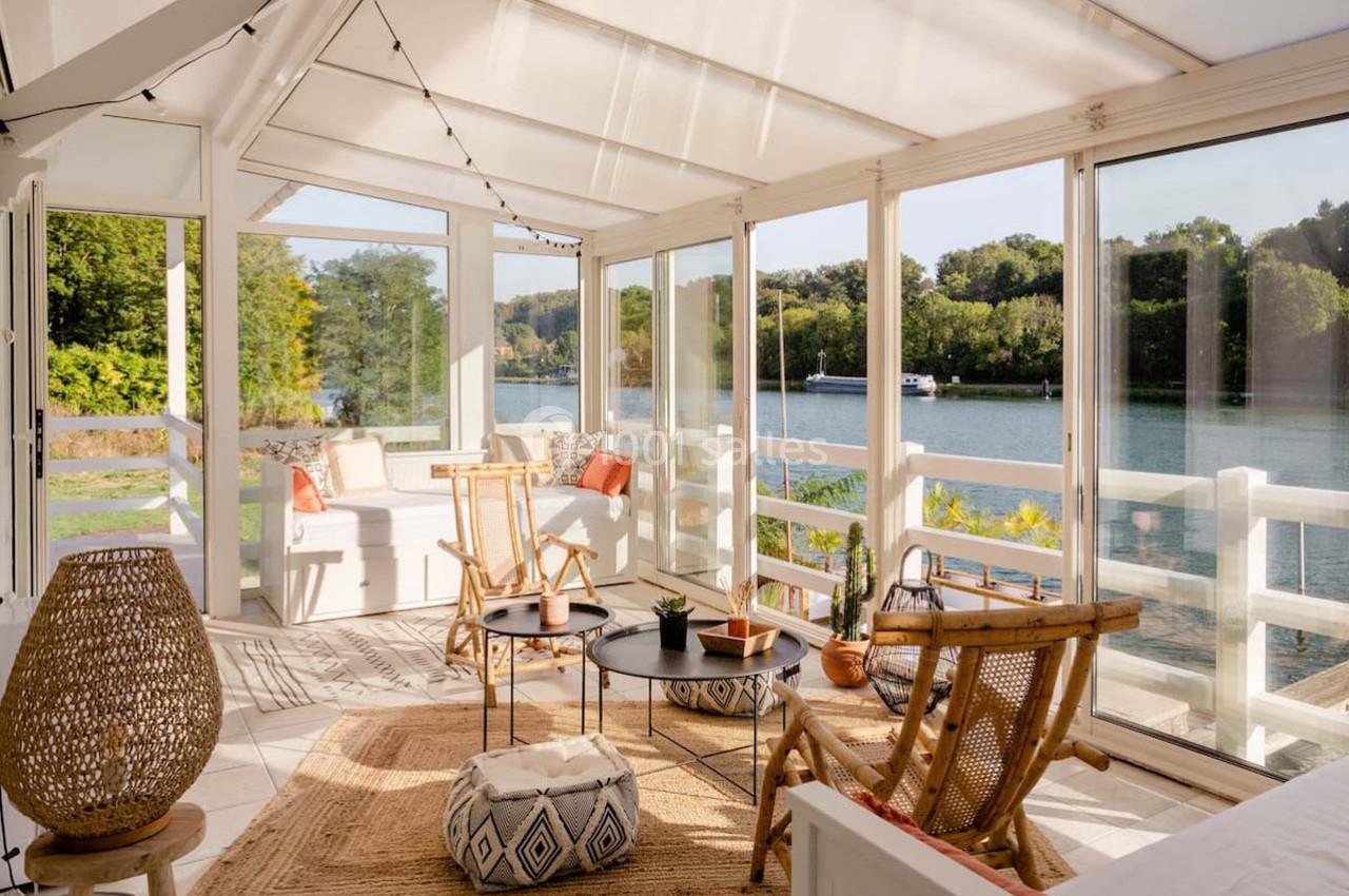 Véranda lumineuse avec mobilier en rotin, tapis beige et vue sur un lac entouré de végétation.