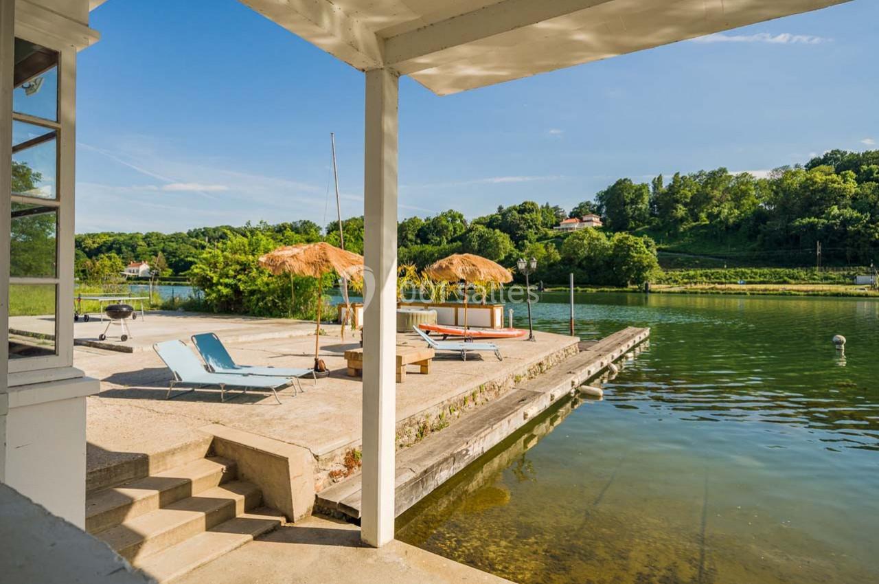 Terrasse au bord d’un lac avec transats, parasols en paille et végétation verdoyante en arrière-plan.