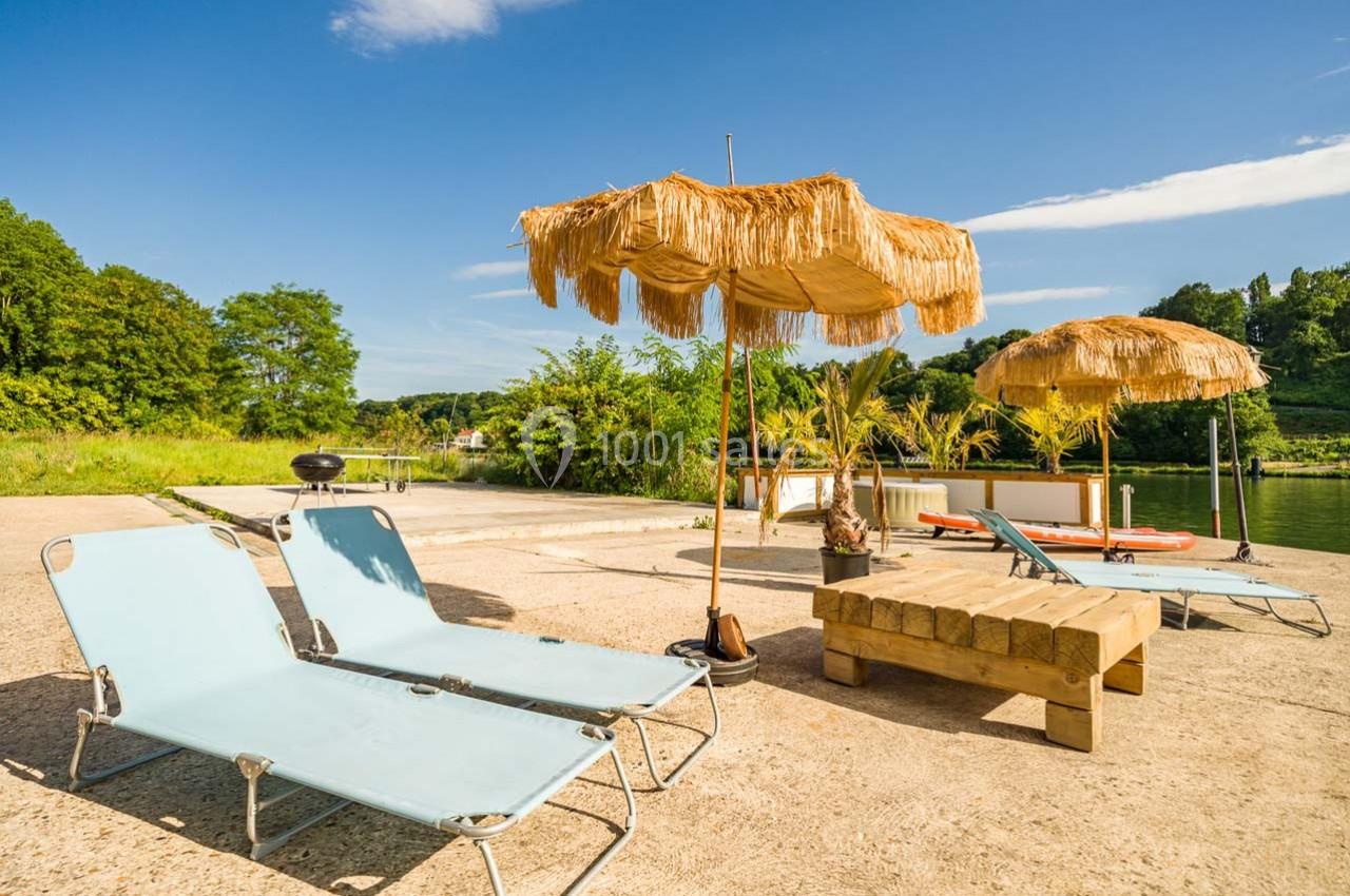 Chaises longues et parasols en paille disposés sur une terrasse ensoleillée près d'un plan d'eau entouré de verdure.