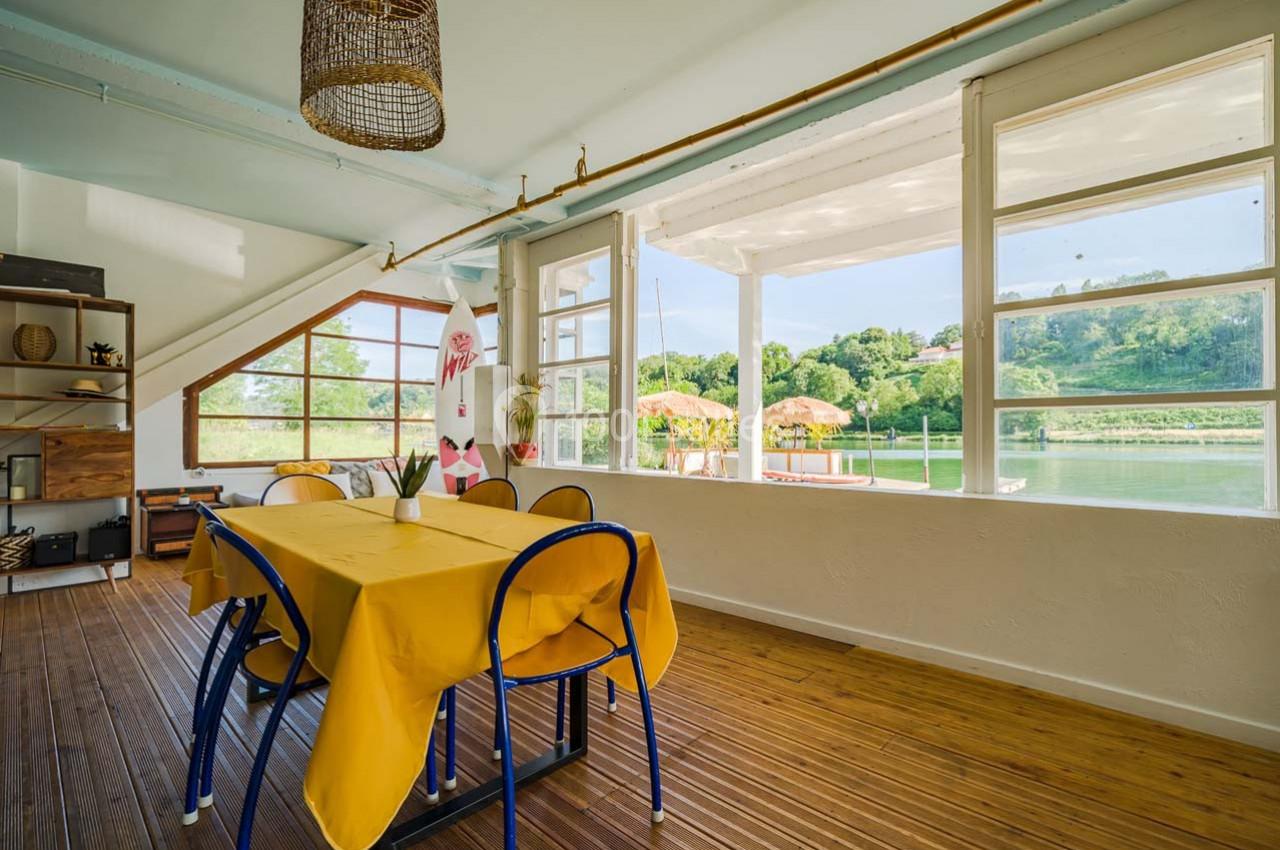 Salle à manger lumineuse avec table jaune, chaises bleues et vue sur une terrasse ensoleillée au bord de l'eau.