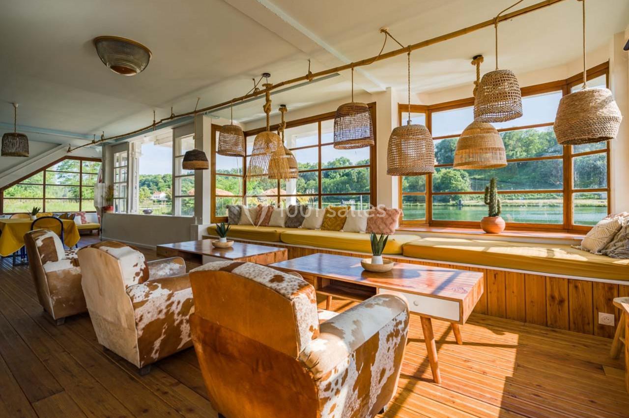Salle lumineuse avec grandes fenêtres, mobilier en bois, fauteuils en tissu et suspensions en osier.