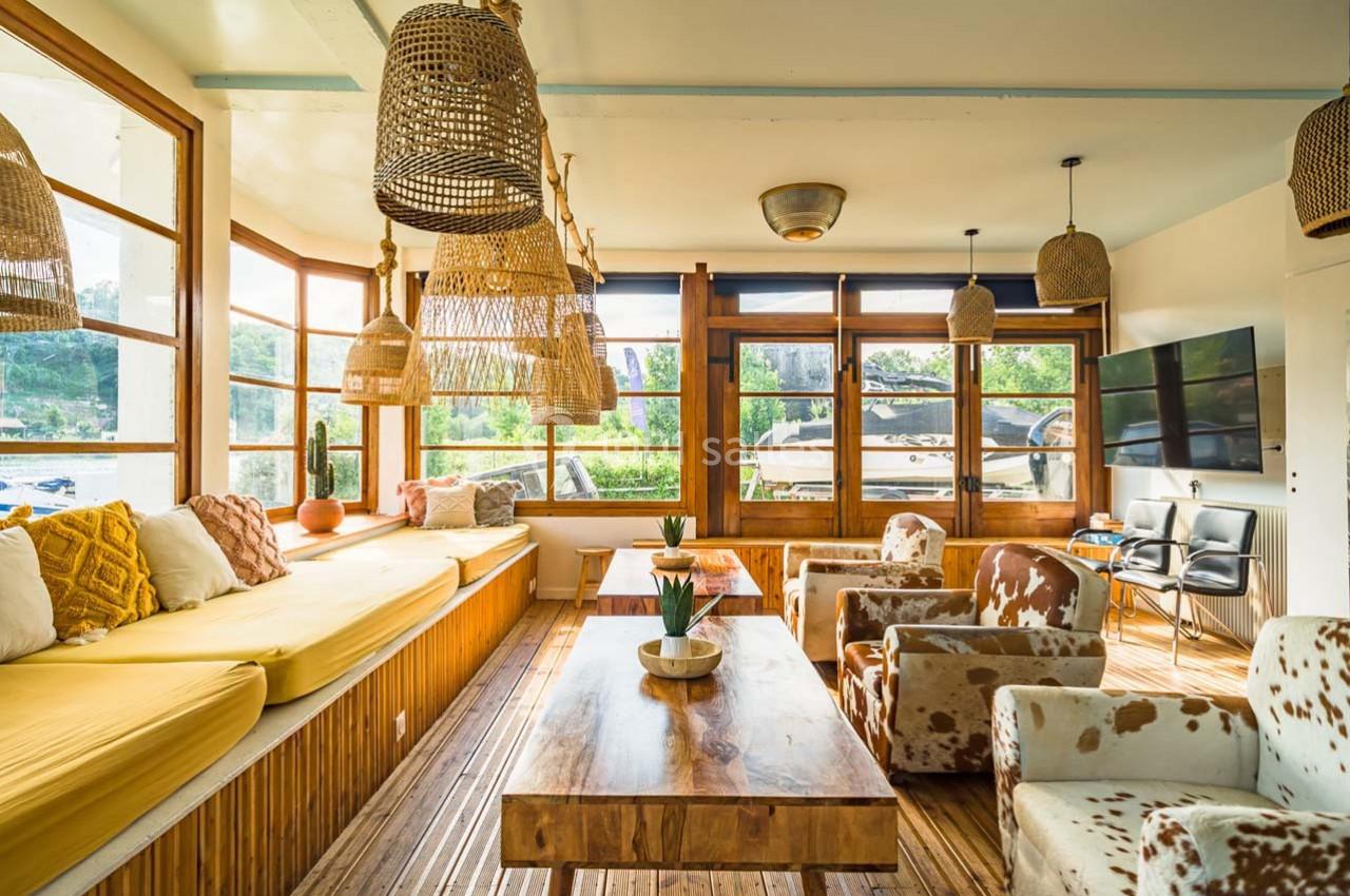 Salon lumineux avec grandes fenêtres, mobilier en bois, banquette jaune, fauteuils imprimés et suspensions en rotin.