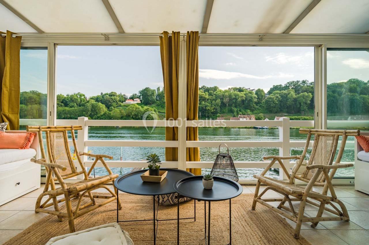 Salon lumineux avec fauteuils en rotin, tables basses noires, tapis en jute et vue sur un lac entouré de verdure.
