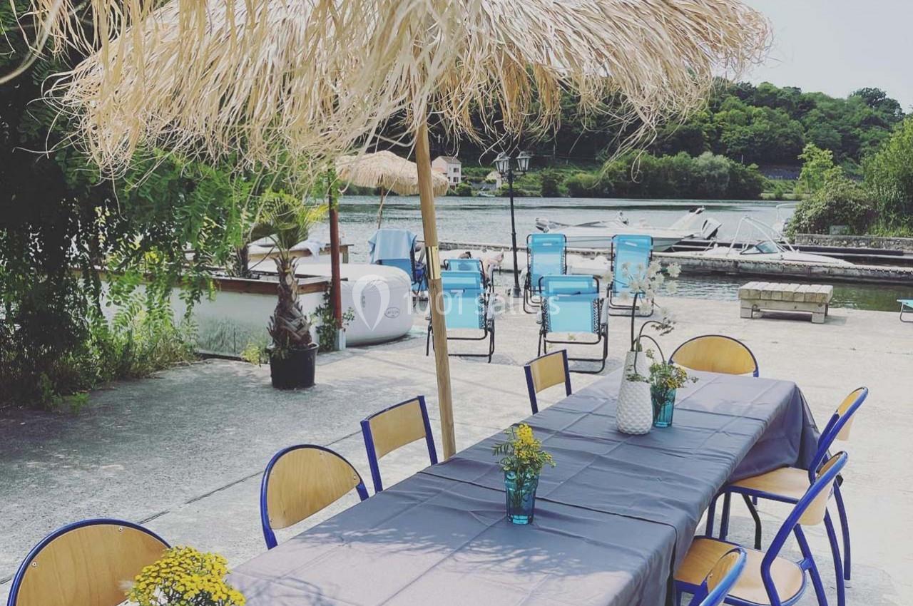 Table dressée sous un parasol en paille, entourée de chaises bleues, avec vue sur un plan d'eau et des bateaux.