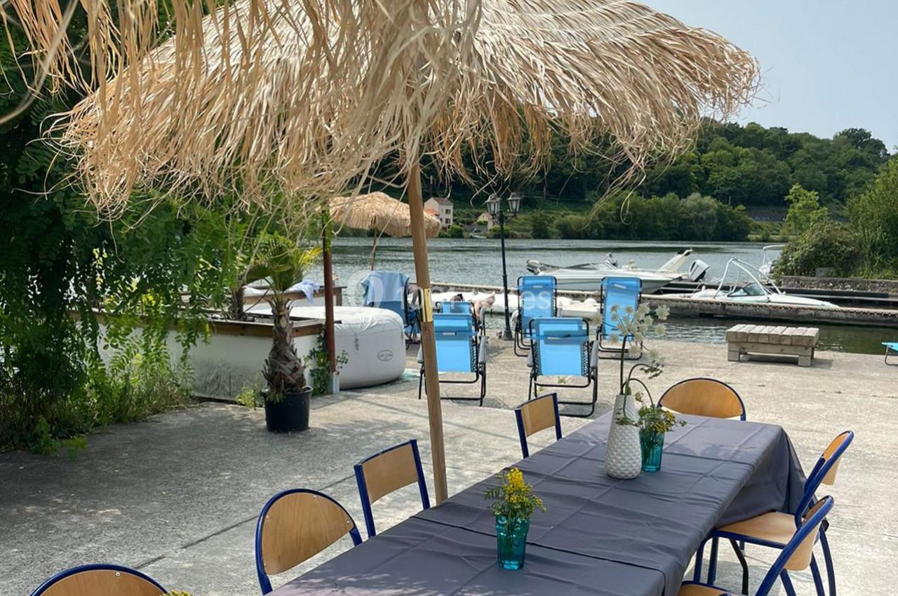 Table dressée avec nappe grise et décorations florales sous un parasol en paille, près d'un quai avec des bateaux.
