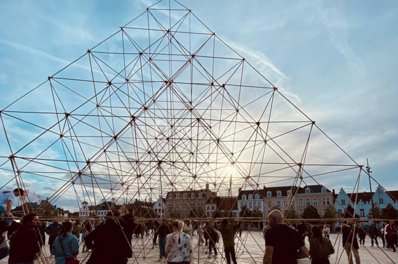 Structure géométrique métallique sur une place publique, entourée de passants sous un ciel partiellement nuageux.