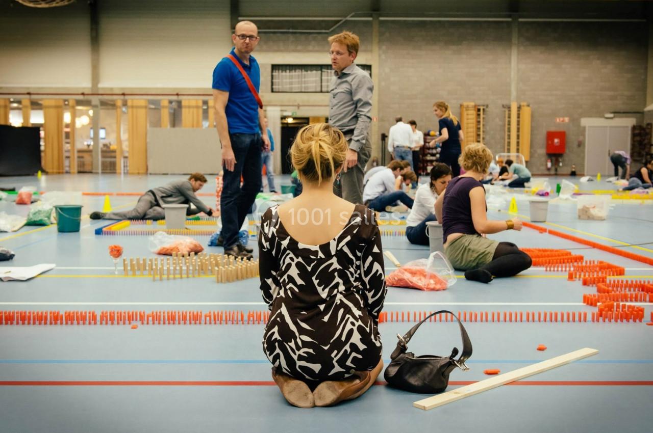 Des personnes assemblent des dominos colorés dans un gymnase, une femme assise au premier plan les observe.