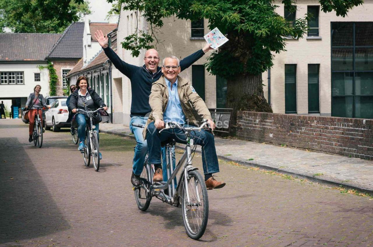Deux hommes souriants sur un tandem, suivis par deux cyclistes, dans une rue pavée bordée de bâtiments.