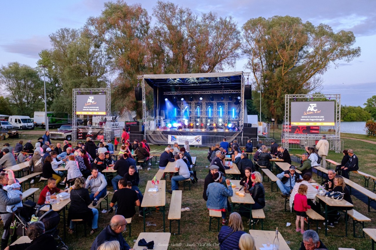 Des personnes assises à des tables en plein air devant une scène éclairée lors d'un événement en soirée.