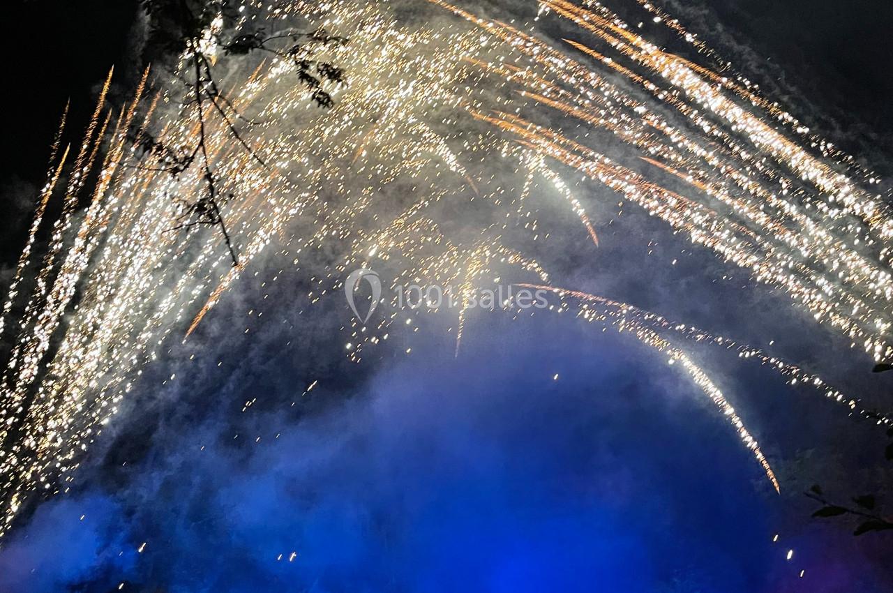 Feu d'artifice illuminant le ciel nocturne avec des éclats lumineux et des projecteurs colorés au sol.