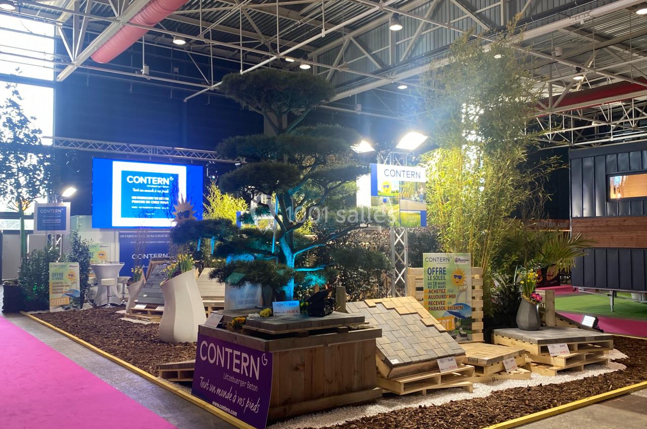 Stand d'exposition avec plantes, matériaux de construction et écrans lumineux dans un hall industriel.