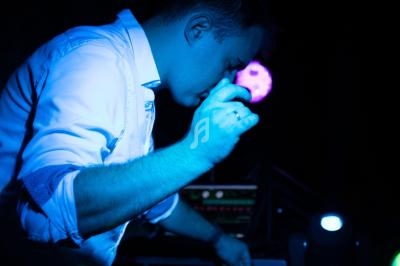 Un DJ en chemise blanche mixe de la musique sur une table de mixage dans une ambiance lumineuse sombre.
