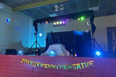 Scène de DJ avec éclairage coloré, deux enceintes et une table recouverte, dans une salle au décor sobre.