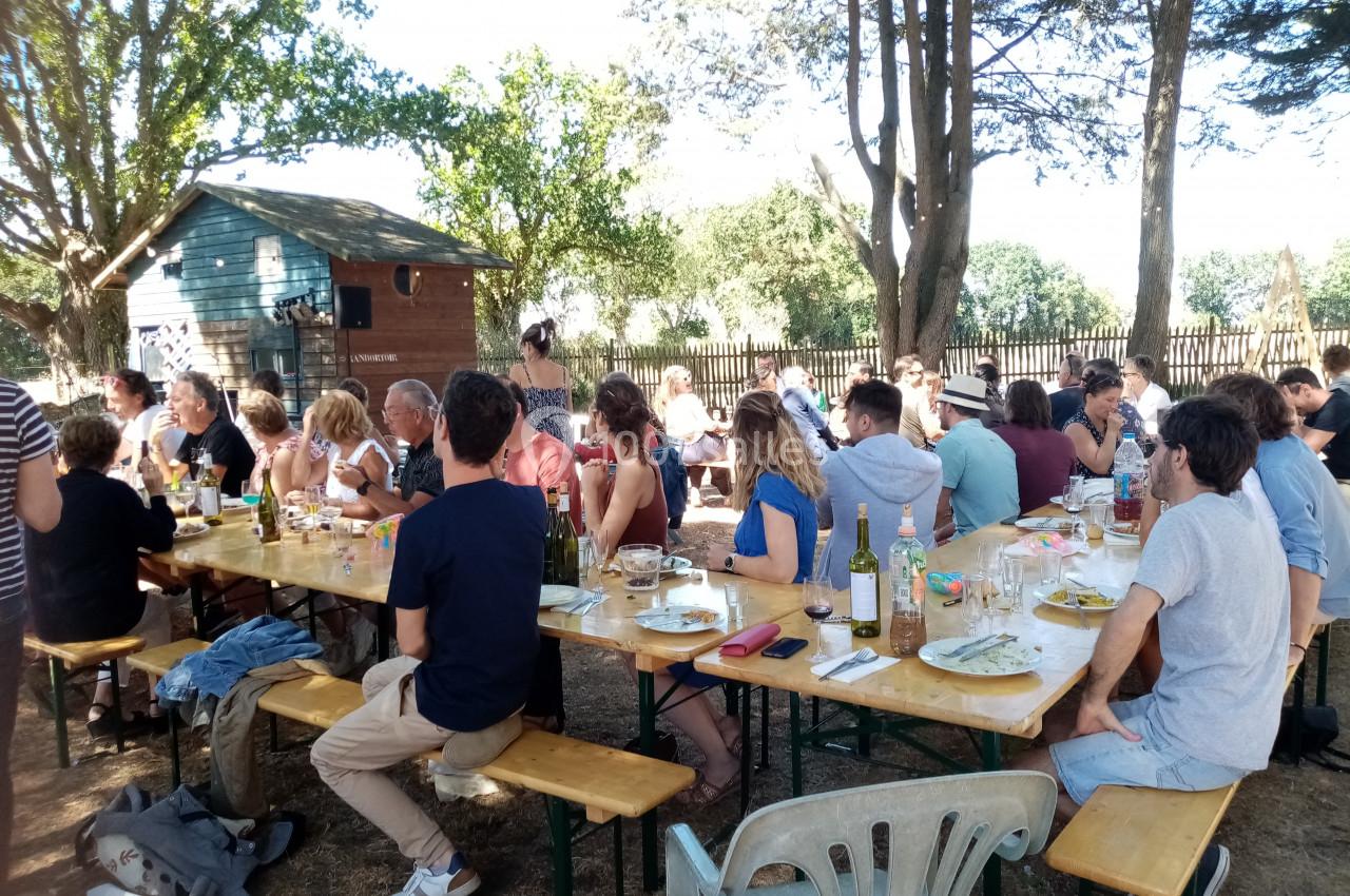 Des personnes assises à des tables en bois partagent un repas en plein air, entourées d'arbres et d'une cabane en arrière…