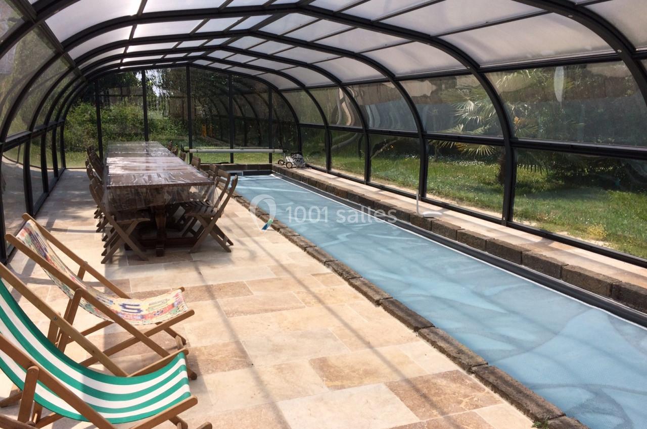 Abri transparent couvrant une piscine étroite, avec une table en bois et des transats alignés sur le côté.
