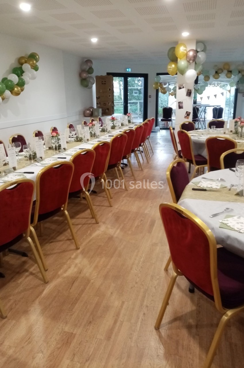 Salle décorée pour un événement avec tables dressées, chaises rouges et ballons suspendus aux murs.