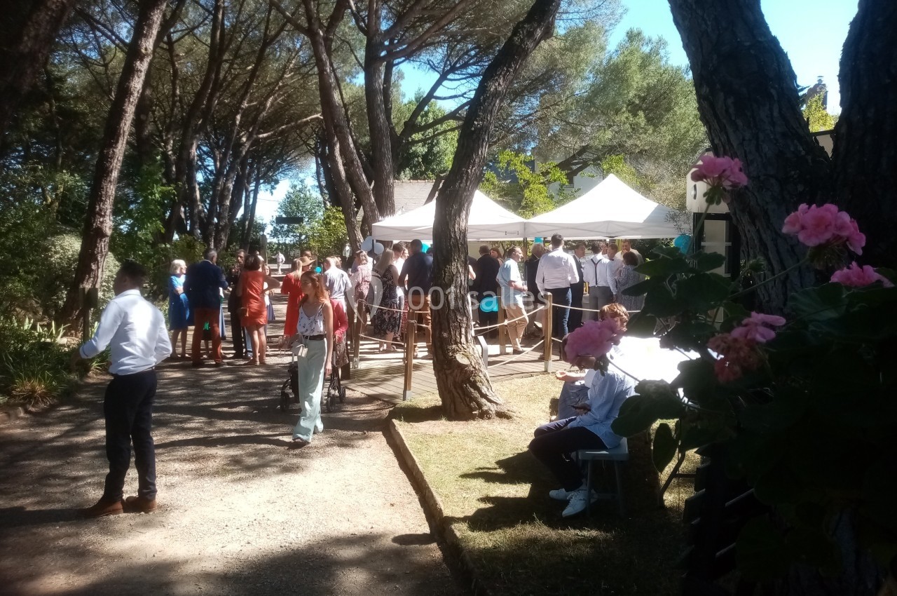 Groupe de personnes rassemblées sous des tentes blanches dans un jardin ombragé bordé d'arbres.