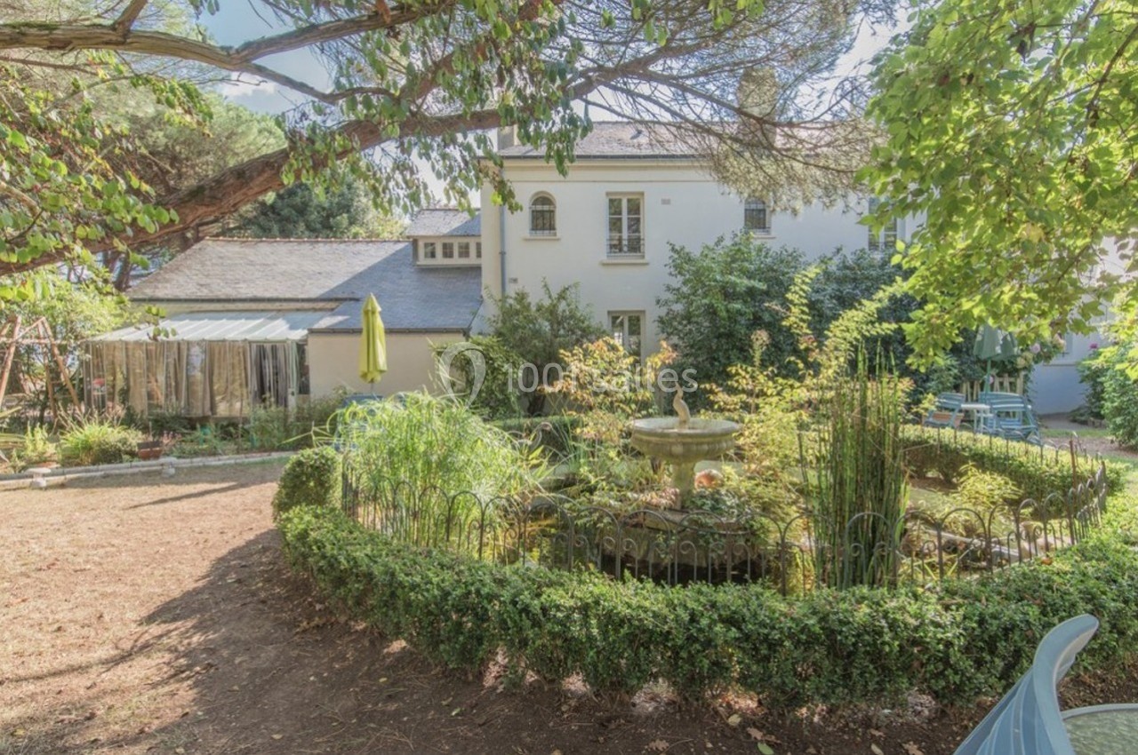 Jardin arboré avec une fontaine centrale entourée de haies, devant une maison blanche à deux étages.