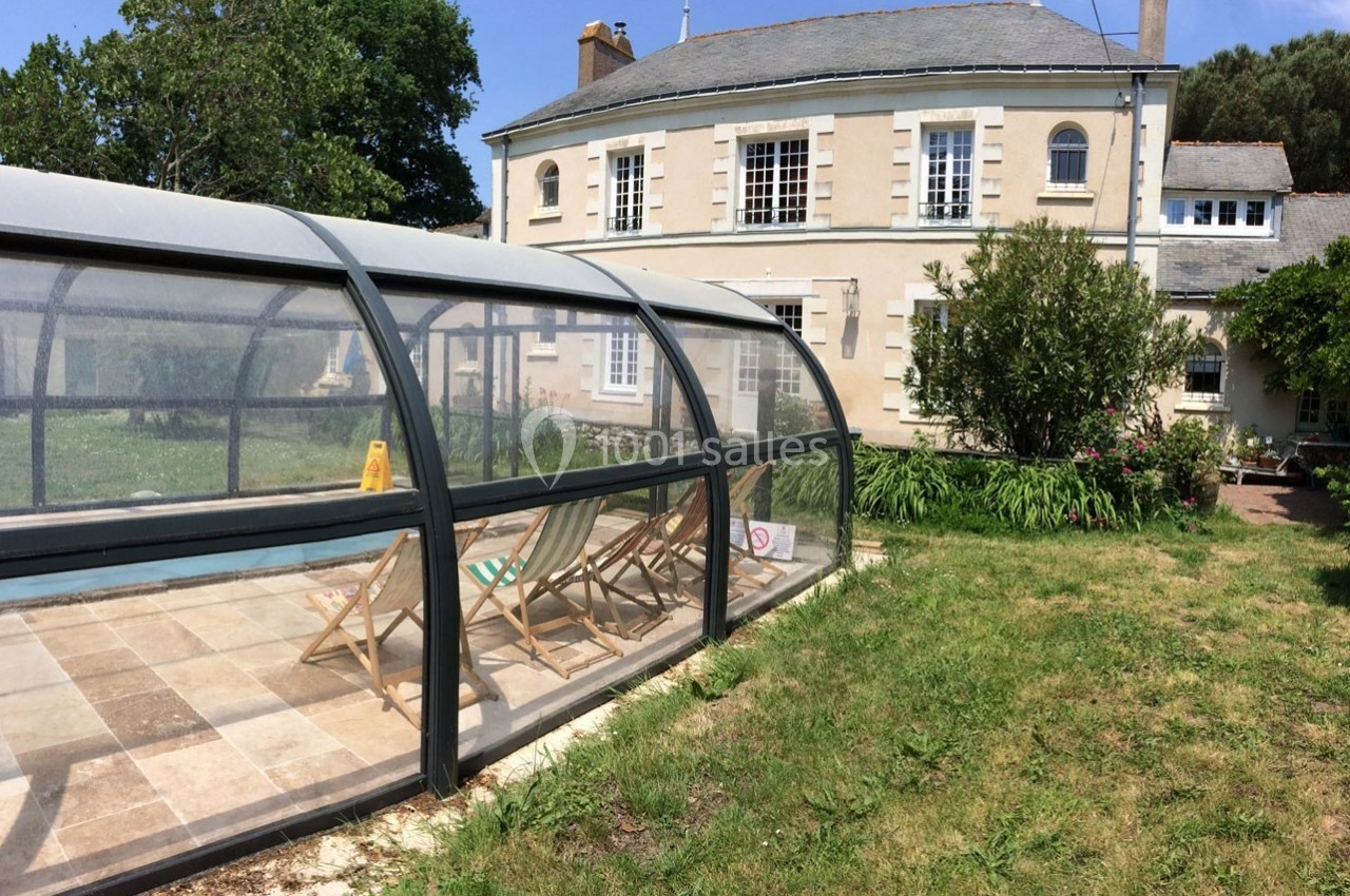 Piscine couverte avec des chaises longues sur une terrasse, devant une grande maison en pierre entourée de verdure.