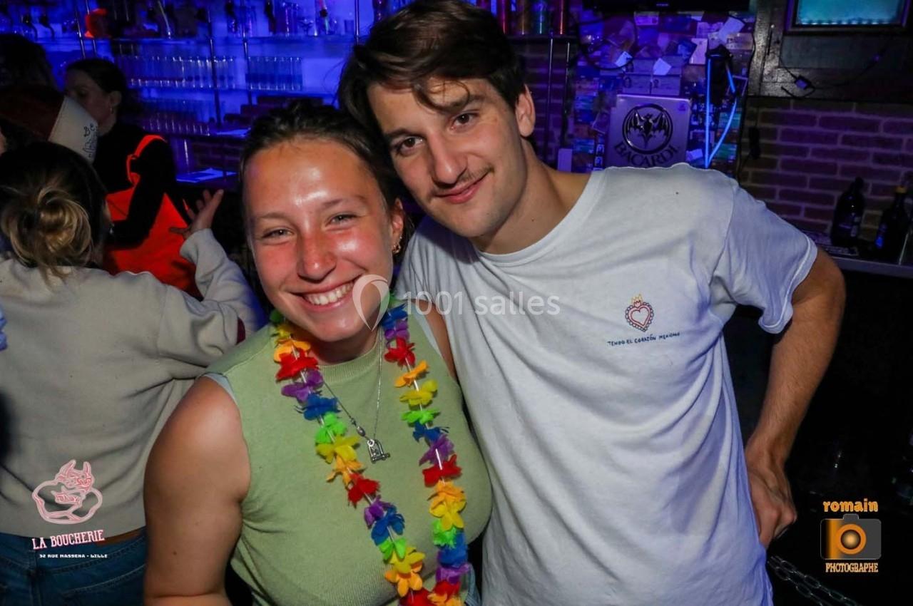 Deux personnes souriantes posent dans un bar, l'une portant un collier de fleurs colorées, avec un fond de lumières bleutées.