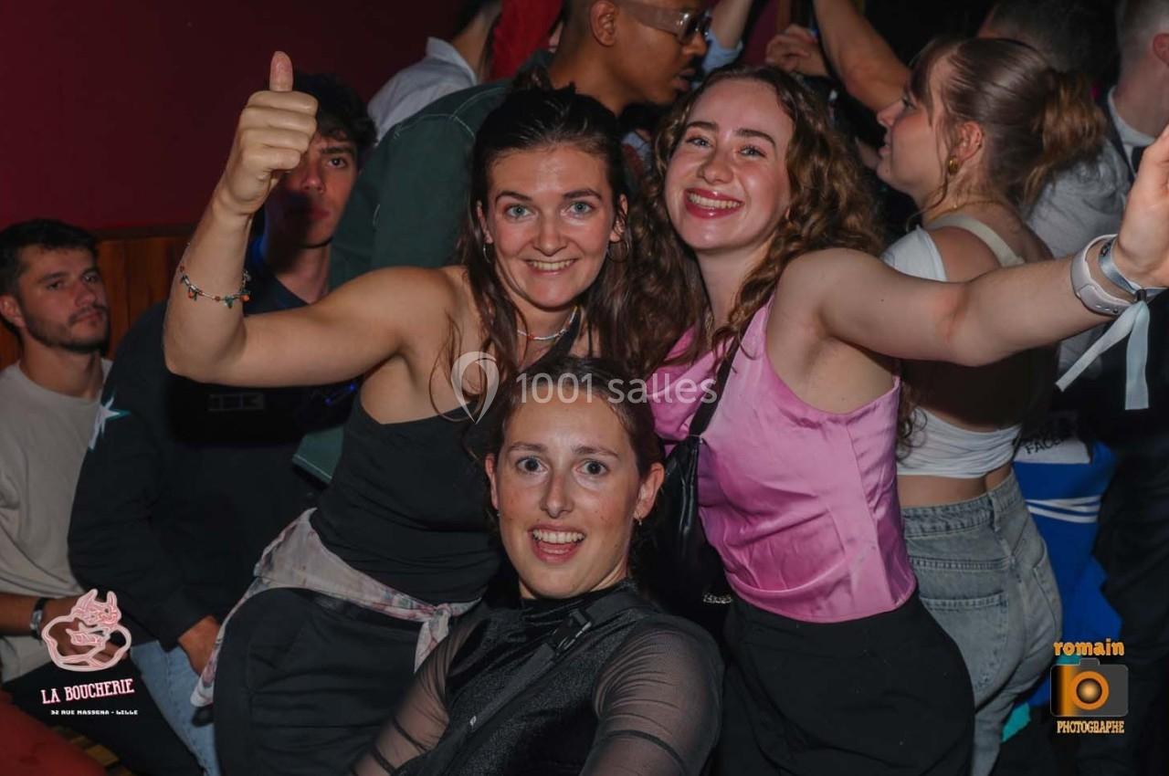 Trois jeunes femmes souriantes posent ensemble dans une ambiance festive, entourées d'autres personnes.
