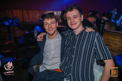 Un homme souriant avec des lunettes de soleil pose avec une femme dans un environnement festif et animé.