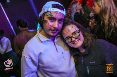 Un homme souriant avec des lunettes de soleil pose avec une femme dans un environnement festif et animé.