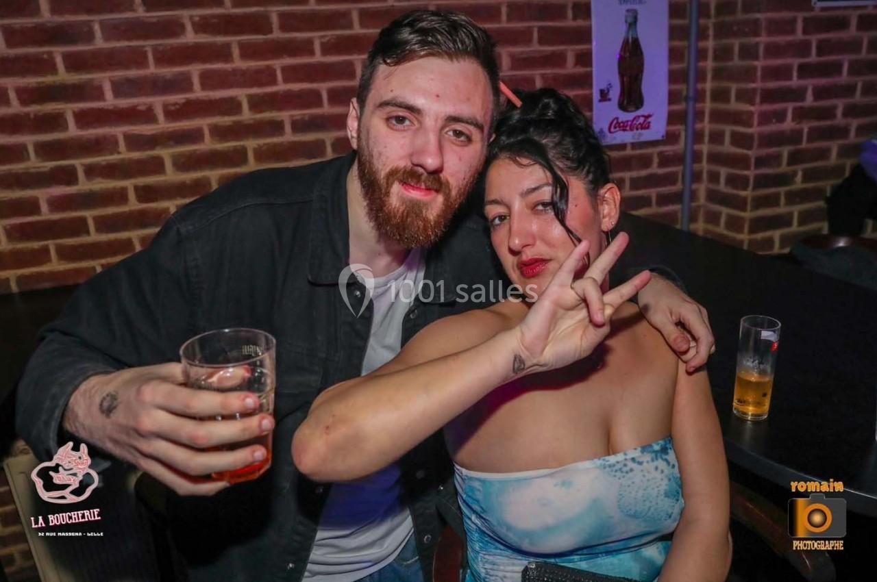 Un homme tenant un verre et une femme faisant un signe de paix posent devant un mur de briques dans un bar.