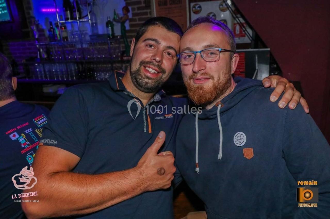 Deux hommes souriants posent côte à côte dans un bar, avec des verres et des bouteilles en arrière-plan.