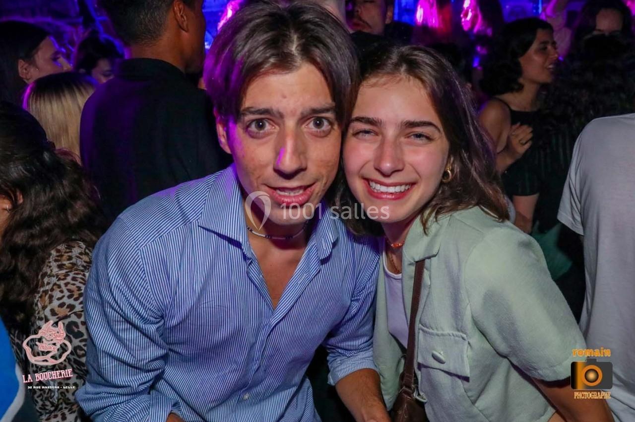 Un homme et une femme souriants posent ensemble dans une ambiance festive avec des lumières colorées en arrière-plan.