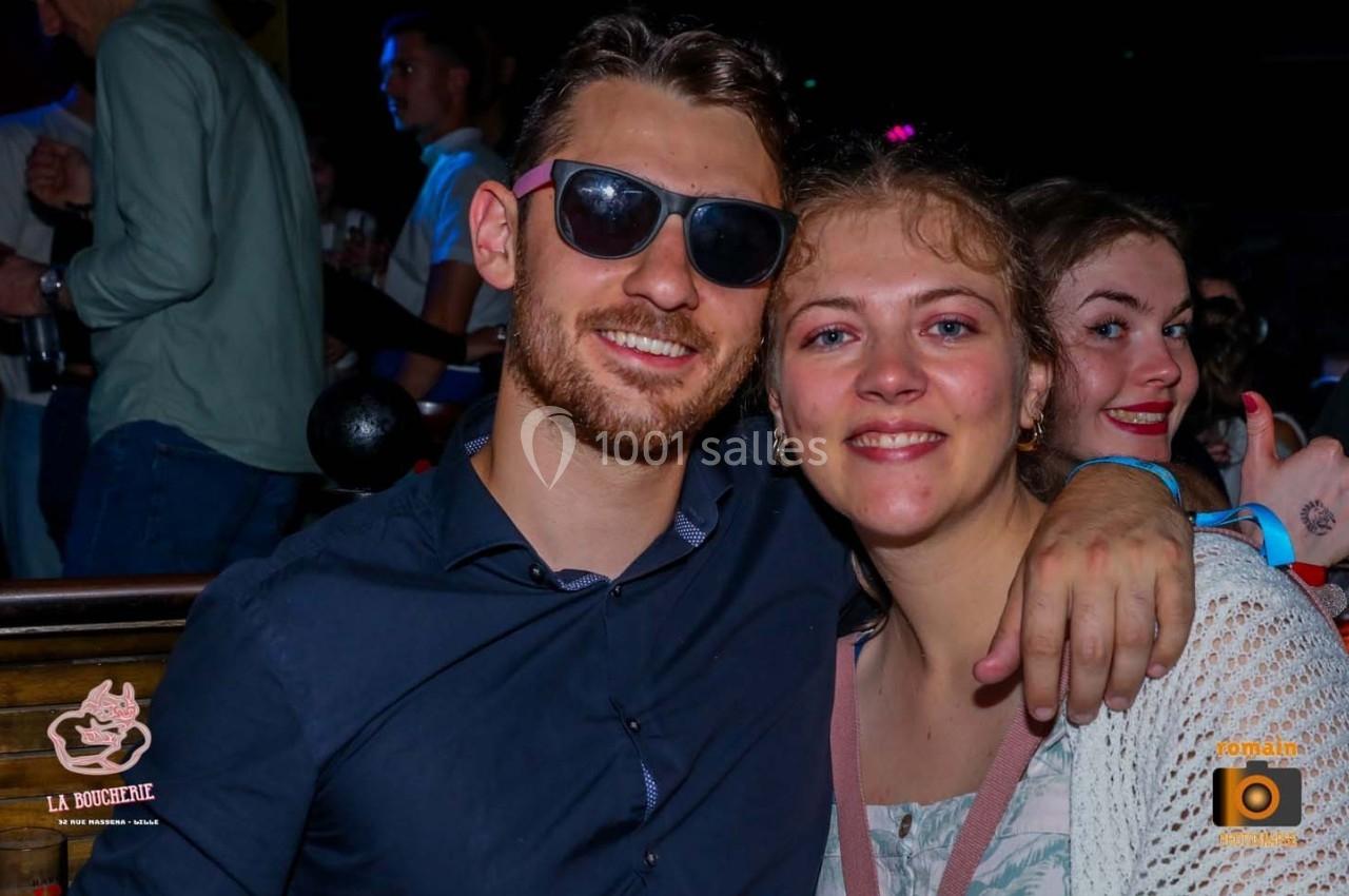 Un homme souriant avec des lunettes de soleil pose avec une femme dans un environnement festif et animé.