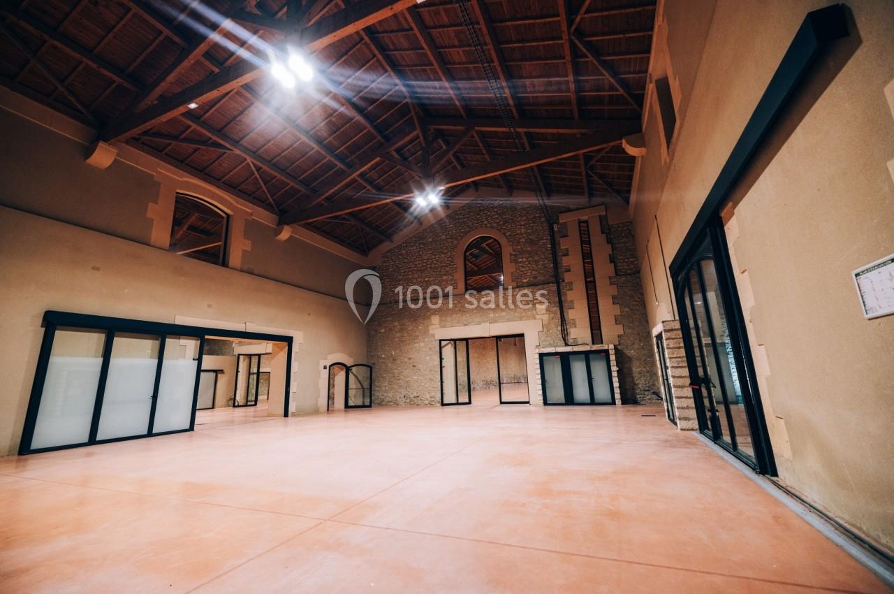 Salle spacieuse avec plafond en bois, murs en pierre et grandes baies vitrées laissant entrer la lumière naturelle.