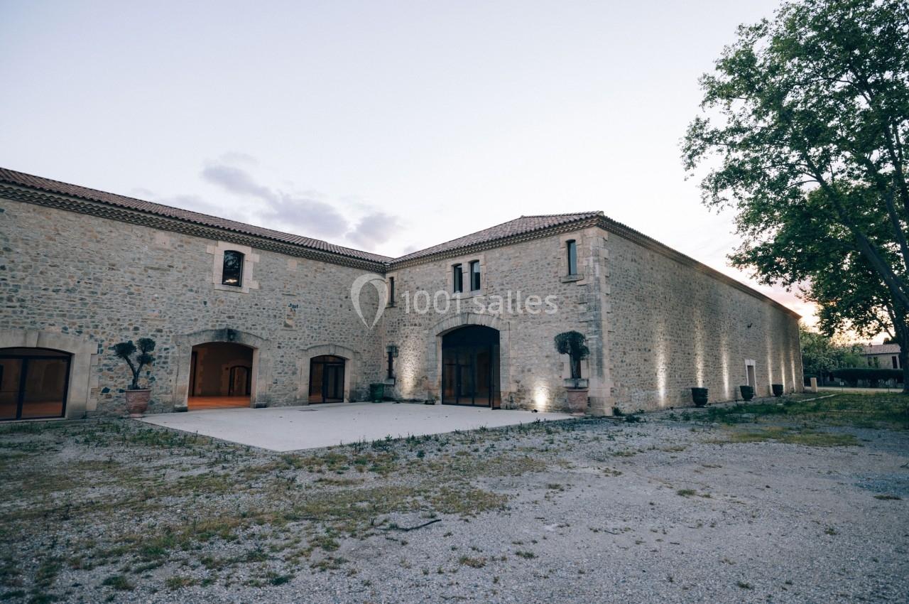Bâtiment en pierre avec grandes ouvertures, éclairé en soirée, entouré d'arbres et d'une cour dégagée.