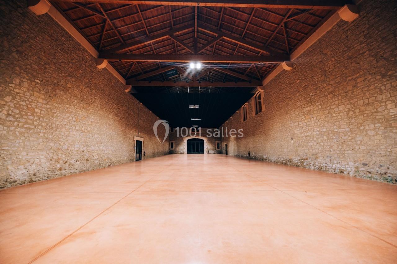 Grande salle vide avec murs en pierre, sol en béton lisse et plafond en bois avec poutres apparentes.
