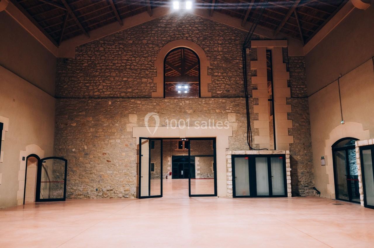 Intérieur d'une grande salle avec murs en pierre, sol en béton lisse, portes vitrées et plafond en bois.