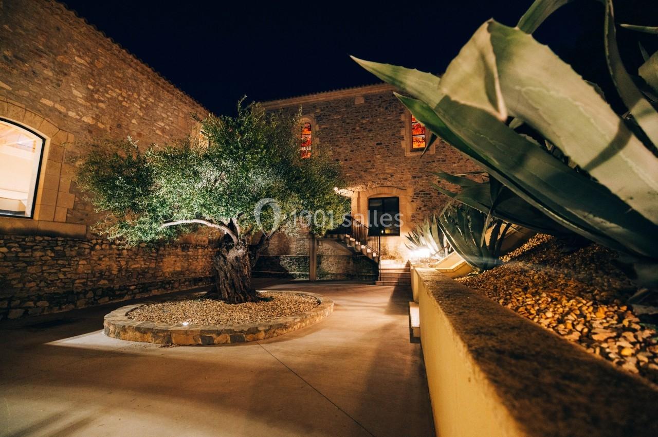 Cour intérieure éclairée de nuit avec un olivier central entouré de pierres et des murs en pierre en arrière-plan.