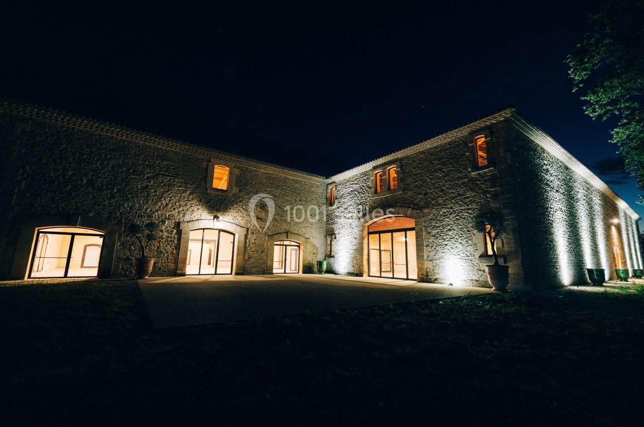 Façade d'un bâtiment en pierre éclairé la nuit, avec de grandes fenêtres et une cour sombre au premier plan.